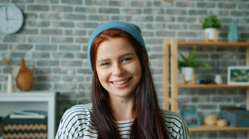 A happy young woman wearing a beanie smiles in a cozy indoor setting with a brick wall backdrop.