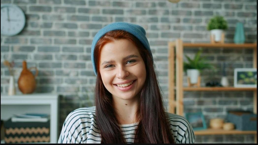 A happy young woman wearing a beanie smiles in a cozy indoor setting with a brick wall backdrop.
